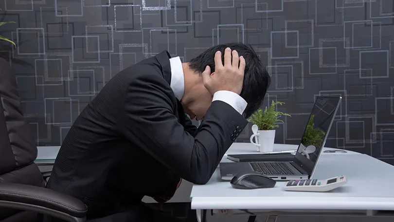 頭を抱えるスーツ姿の男性がデスクで悩んでいる様子。